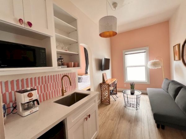 A cozy small apartment features a kitchen with a sink and coffee maker, a living area with a couch, TV, and stylish decor, and a pink accent wall.