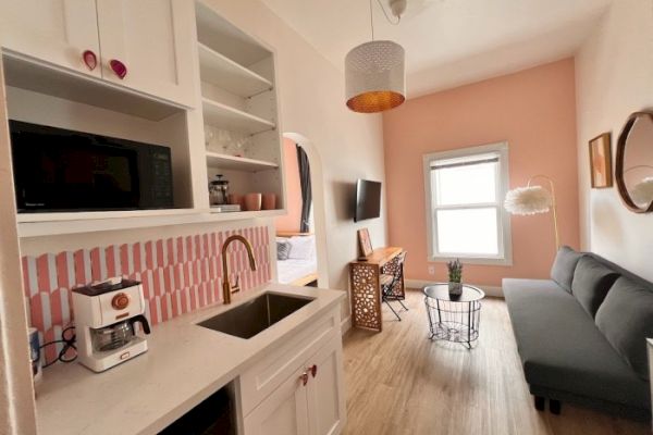 A cozy room with a kitchenette, pink and white decor, a gray sofa, flat-screen TV, hanging light, and a window with natural light coming in.