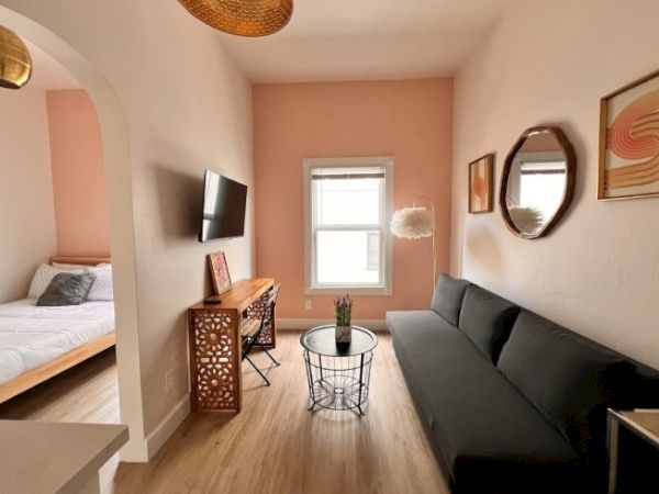 The image shows a cozy living space with a couch, coffee table, wall-mounted TV, and a connected bedroom with a bed.