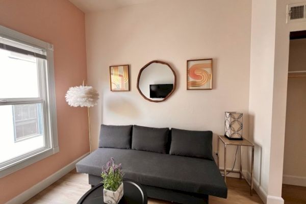 A cozy living room with a gray couch, round mirror, framed art, floral lamp, and potted plant on a round coffee table near a window with peach walls.