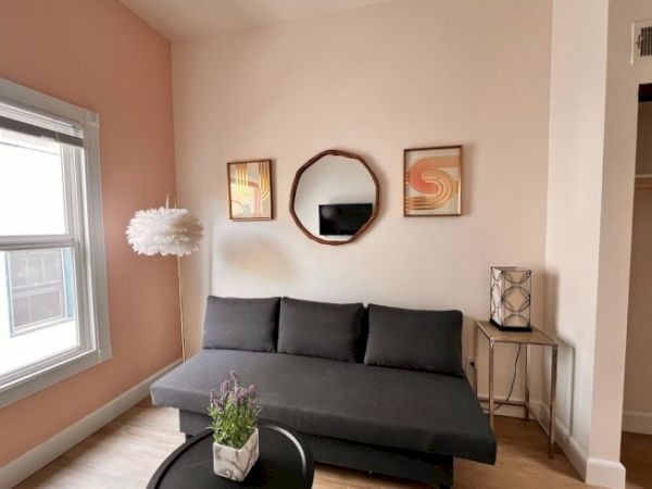 A cozy living room with a gray couch, round mirror, framed art, floral lamp, and potted plant on a round coffee table near a window with peach walls.