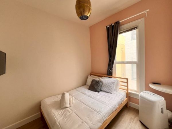 A small bedroom with a double bed, pillows, a rolled towel, a hanging lamp, a window with curtains, a TV, and an air conditioner.