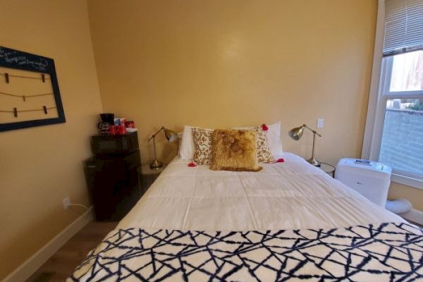 A neatly made bed with white sheets and decorative cushions sits in a cozy bedroom with beige walls, bedside tables, lamps, and a patterned blanket.