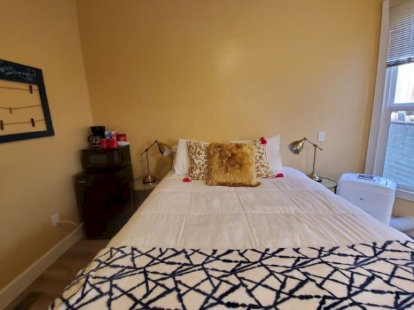 A neatly made bed with white sheets and decorative cushions sits in a cozy bedroom with beige walls, bedside tables, lamps, and a patterned blanket.