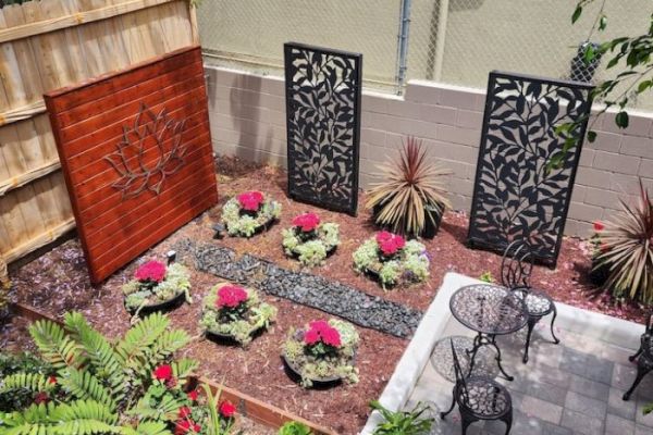 A small garden with decorative panels, potted plants with red flowers, a gravel path, and an outdoor seating area with two chairs and a table.