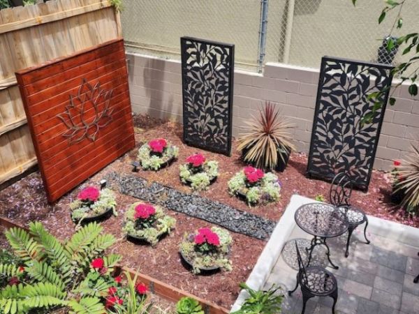 A small garden with decorative panels, potted plants with red flowers, a gravel path, and an outdoor seating area with two chairs and a table.
