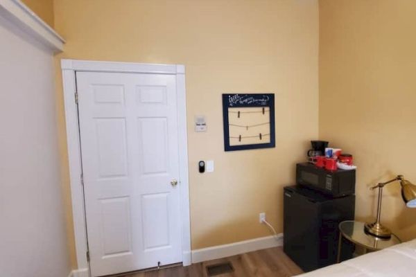 A small room with a yellow wall, a white door, a black mini fridge, coffee supplies on top, a lamp, and a wall-mounted coat rack.