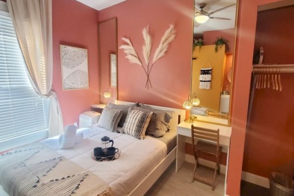 A cozy bedroom features a neatly made bed with pillows, a small tray, a desk with a mirror, and decorative elements on a pinkish wall.