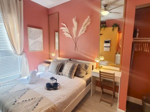 A cozy bedroom features a neatly made bed with pillows, a small tray, a desk with a mirror, and decorative elements on a pinkish wall.