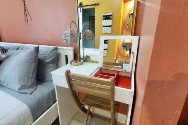 A cozy bedroom features a wooden chair at a white vanity desk with a mirror, a lamp, and a bed with gray pillows and a coral wall.