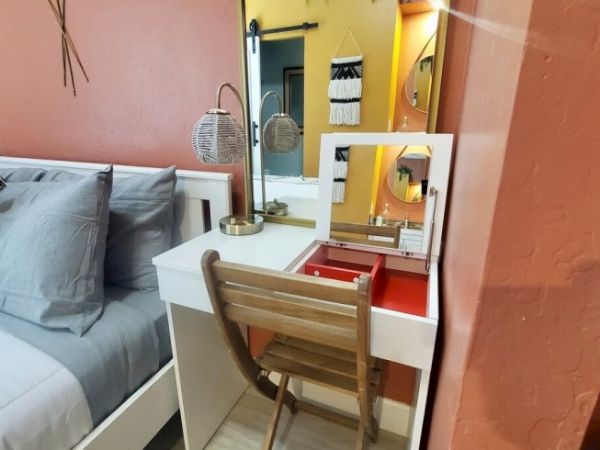 A cozy bedroom features a wooden chair at a white vanity desk with a mirror, a lamp, and a bed with gray pillows and a coral wall.