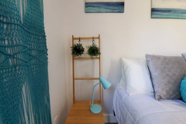 The image shows a bedroom with a bed, a nightstand with a blue lamp, wall art, hanging plants, and a teal macrame wall hanging.