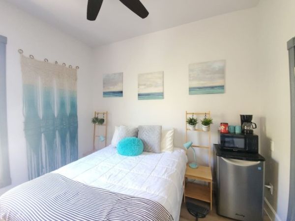 A cozy bedroom with a bed, teal pillows, wall art, hanging plants, a coffee maker, a microwave, and a mini-fridge. The room has a serene vibe.