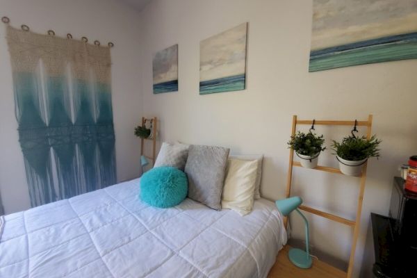 A cozy bedroom with a white bedspread, decorative pillows, wall art, hanging planters, and a knitted wall piece, exuding a serene, beachy vibe.