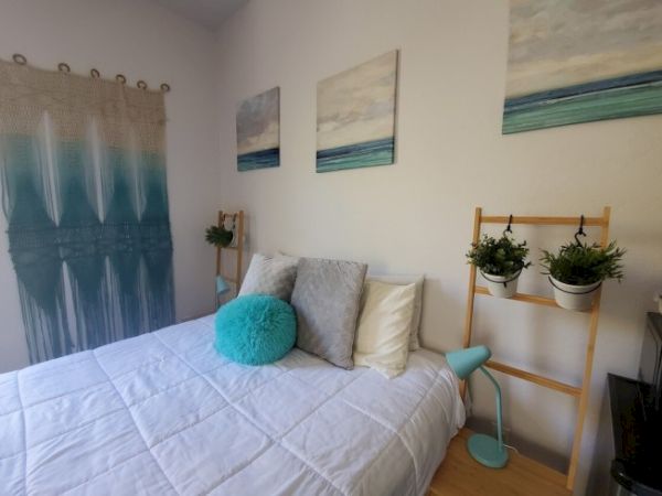 A cozy bedroom with a white bedspread, decorative pillows, wall art, hanging planters, and a knitted wall piece, exuding a serene, beachy vibe.